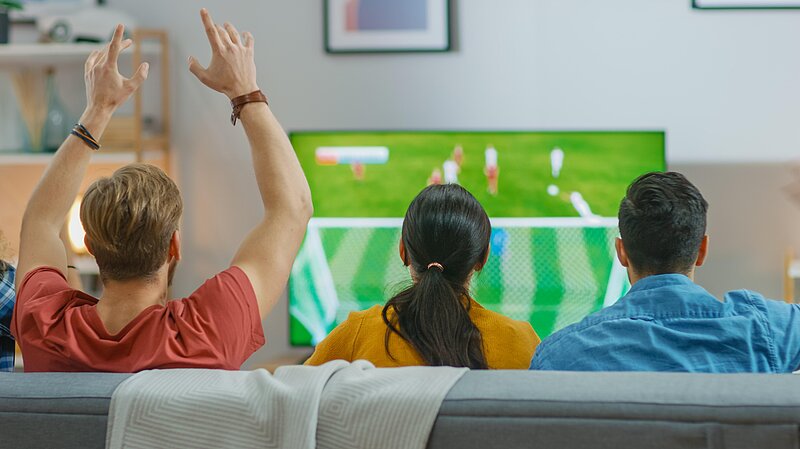 Photograph showing three people from behind, the person on the left has his hands in the air, the people are watching a football match on TV, the match is out of focus.