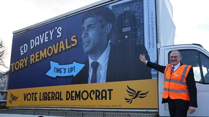 Photo of Lib Dem leader Ed Davey in front of poster van. Poster van reads "Ed Davey's Tory Removals" "Vote Liberal Democrat" A large arrow with the words "Time's up" points to an image or Rishi Sunak outside 10 Downing Street.
