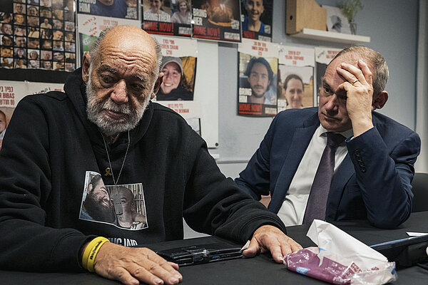 An visibly upset elderly man wearing a black hoody with photos of his two sons on the front, his sons were  kidnapped by Hamas on 7 October 2023. Next to him sits Ed Davey, holding his head with one hand while listening to the man. The wall behind the two men shows posters of other hostages.
