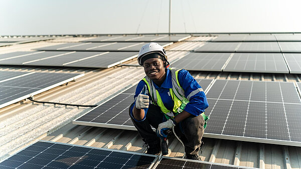 Photo a man in a blue shirt and a yellow safety helmet crouches on a roof of a building that has solar panels on it.