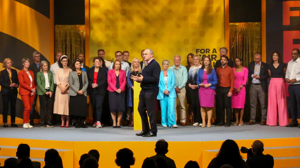 Ed Davey on stage at Liberal Democrat conference. Behind him are around 30 Liberal Democrat MPs and candidates, to the left is the Lib Dem stunt from the Somerton and Frome by-election: a blue cannon with the words "Get these clowns out of Number 10!", to the right is the stunt from the 2023 local elections: a large blue clock with the words "Time's up for Rishi Sunak". In the foreground the conference audience can be seen.