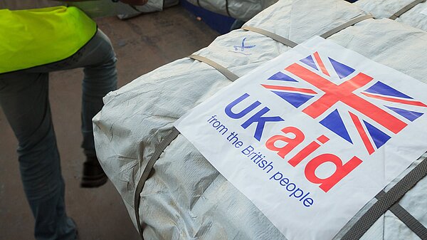 Two men are moving parcels wrapped in white plastic. In the foreground, a label on one package is visible reading "UK Aid from the British people" and a union flag. Image - Dfid CCBY-ND2.0