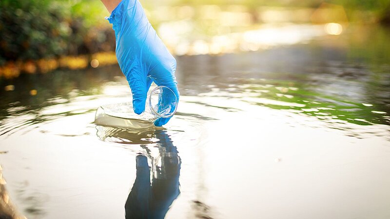 A hand in a plastic glove is using a flask to collect a sample from standing water