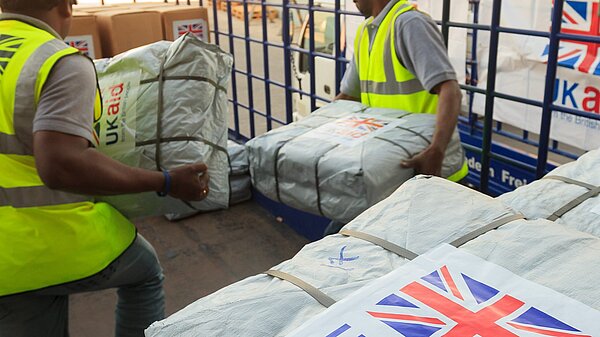 Two men are moving parcels wrapped in white plastic. In the foreground, a label on one package is visible reading "UK Aid from the British people" and a union flag. Image - Dfid CCBY-ND2.0