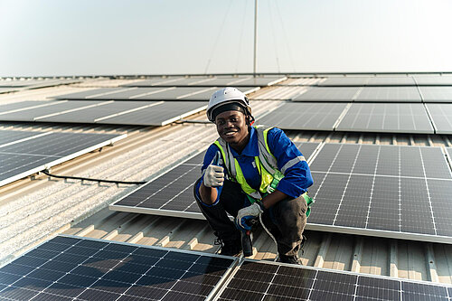 Photo a man in a blue shirt and a yellow safety helmet crouches on a roof of a building that has solar panels on it.