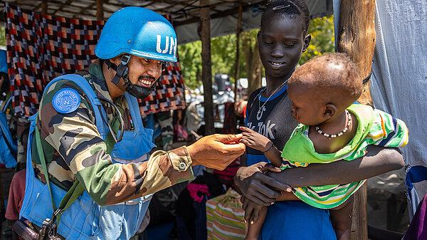 A man in blue UN body armour and helmet plays with a young child being held by a woman