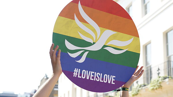 Young person holding a round sign over their head with a rainbow-striped, pride flag design overlaid with the Liberal Democrat bird of liberty emblem and #LoveIsLove
