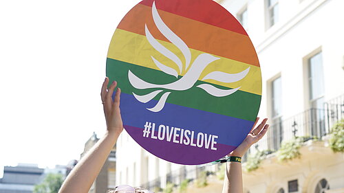 Young person holding a round sign over their head with a rainbow-striped, pride flag design overlaid with the Liberal Democrat bird of liberty emblem and #LoveIsLove